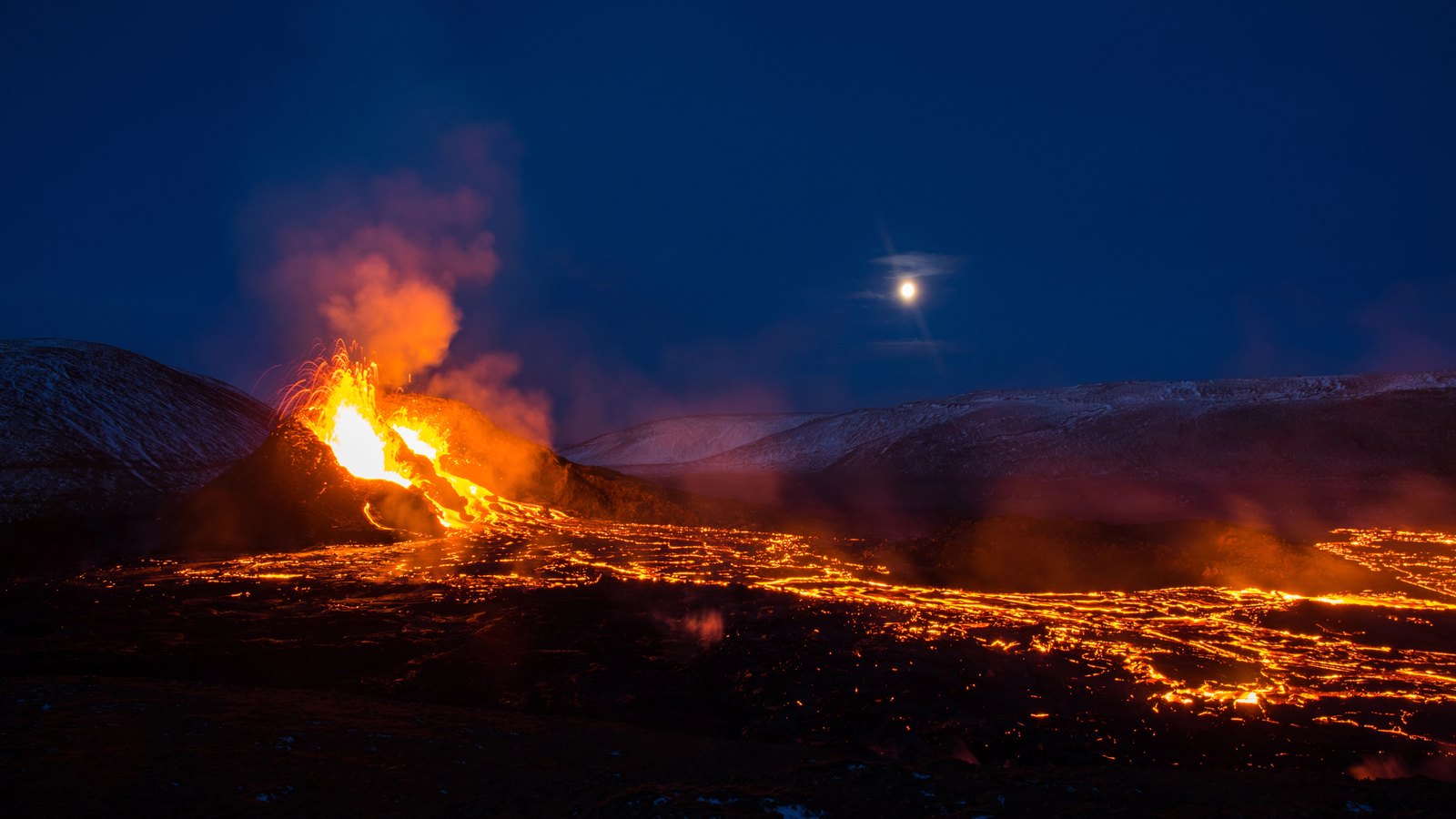 Lavafontäne an einem kleinen Kegel, davor ein weit ausgedehntes glühendes Lavafeld in der Dämmerung